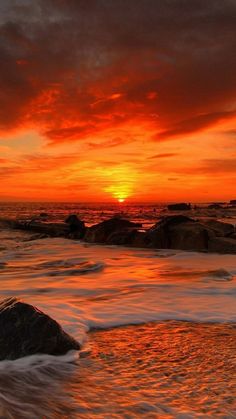 the sun is setting over the ocean with rocks in the foreground and waves on the shore