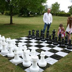 two children playing chess on a giant checker board in the grass with an older man watching