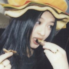 a woman with long black hair wearing a yellow hat and holding a piece of food in her mouth