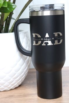 a black travel mug with the word dad on it next to a potted plant