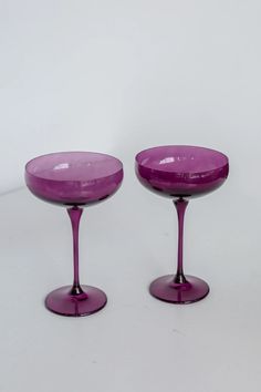 two purple wine glasses sitting next to each other on a white surface with no one around them