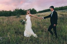 a bride and groom holding hands in the middle of a field at sunset with pink clouds