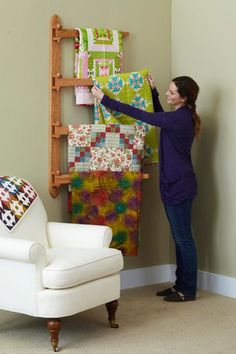 a woman standing in front of a wall hanging quilts on a wooden ladder next to a white chair