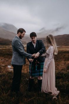 two men and a woman standing in the middle of a field with mountains behind them