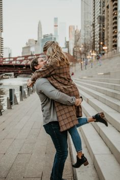 a man and woman are hugging on the steps in front of a river with tall buildings