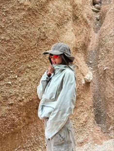 a woman standing in front of a rock wall wearing sunglasses and a hat with her hands on her hips