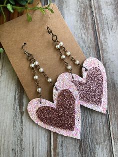 two heart shaped earrings with pearls hanging from them on a piece of paper next to a plant