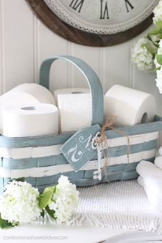 a basket filled with toilet paper on top of a table next to flowers and a clock