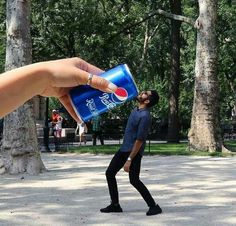 a hand holding a can of pepsi in front of a man walking across a park