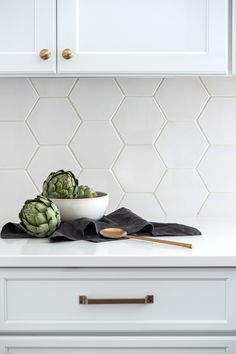 two artichokes in a white bowl on a kitchen counter with gold spoons