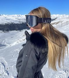 a woman wearing ski goggles standing on top of a snow covered slope with her hair blowing in the wind