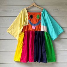 a rainbow colored dress hanging on a wooden wall next to a hanger with an orange, yellow, green, blue, and pink shirt