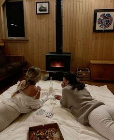 two women sitting on a bed playing cards in front of an open fire place,