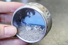 a person is holding a compact camera lens with the reflection of a beach and palm trees in it