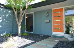 an orange front door on a blue house with gravel and trees in the foreground