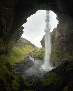 a waterfall coming out of the side of a cave into a river surrounded by lush green grass