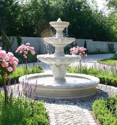 a white fountain surrounded by pink flowers and greenery in the middle of a garden