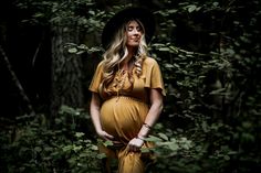 a pregnant woman standing in the woods wearing a yellow dress and black hat with her hands on her hips