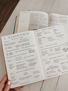 an open book with writing on it sitting on top of a wooden table next to a person's hand