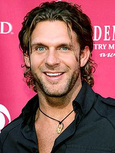 a man with long hair and blue eyes smiles at the camera while standing in front of a pink backdrop