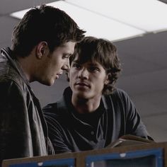 two young men standing next to each other looking at something on the table in front of them