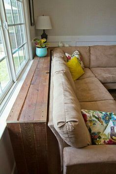 a couch sitting in front of a window next to a wooden table with pillows on it