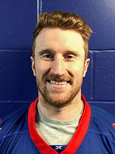 a man with a beard wearing a blue jersey and smiling at the camera while standing in front of a blue wall