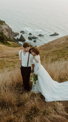 a bride and groom standing in the grass by the ocean