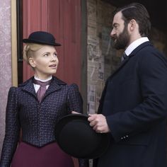 a man standing next to a woman with a hat on her head and wearing a suit