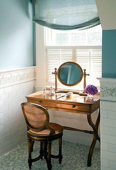 a desk with a mirror and chair in front of a window next to a wall