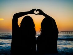 two women standing in front of the ocean making a heart shape with their hands at sunset