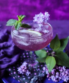 a purple drink with ice and mint garnish on a blue plate next to flowers