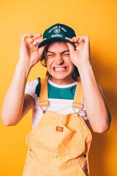 a woman in overalls and a hat covering her eyes with one hand while standing against a yellow wall