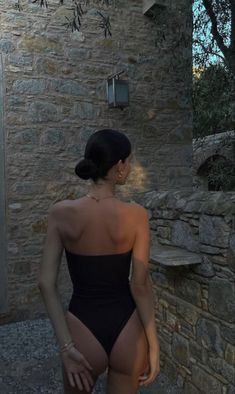 a woman in a black swimsuit standing next to a stone wall and looking into the distance