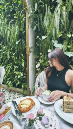 a woman sitting at a table with plates of food on it and plants in the background