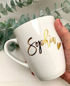 a hand holding a white coffee mug with gold lettering