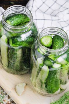 two jars filled with pickles sitting on top of a cutting board next to cucumbers