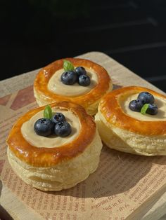 three pastries with blueberries and cream on top sit on a wooden cutting board