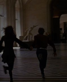 two people are running in the middle of a room with statues on either side of them