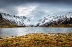 the mountains are covered in snow and green mossy grass near a body of water