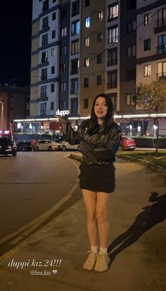 a woman standing on the side of a road in front of tall buildings at night