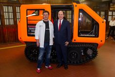 two men in suits standing next to an orange vehicle that is shaped like a tractor