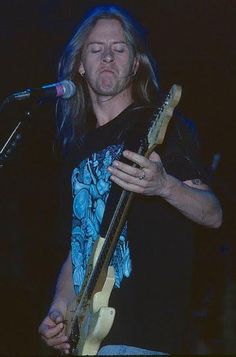 a man with long hair holding a guitar in front of a microphone and singing into a mic