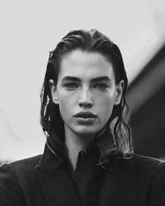 a black and white photo of a young woman with long hair wearing a coat looking at the camera