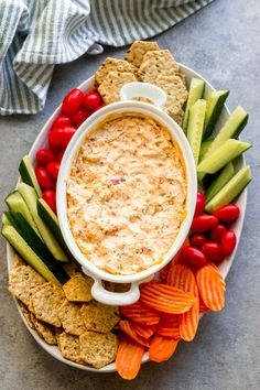 a bowl of dip surrounded by vegetables and crackers on a platter with tomatoes, cucumbers, celery, carrots