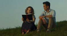 a man and woman sitting in the grass while reading a book, one is looking up at the sky