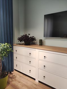 a white dresser with drawers and a television mounted on it's wall next to a potted plant