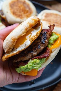 a hand holding a bacon, egg and avocado sandwich with eggs on the side