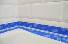 the corner of a bathtub with blue tape on it's edge and white tiles