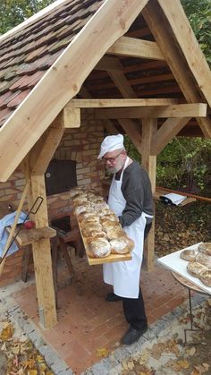 a man in an apron holding a large loaf of bread under a wooden shelter with brick walls